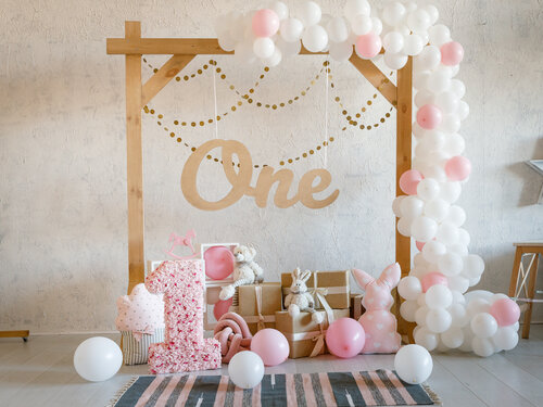 Birthday decorations with wooden arch, gifts, toys, balloons, garland and figure 1 for little baby party on a white wall background.